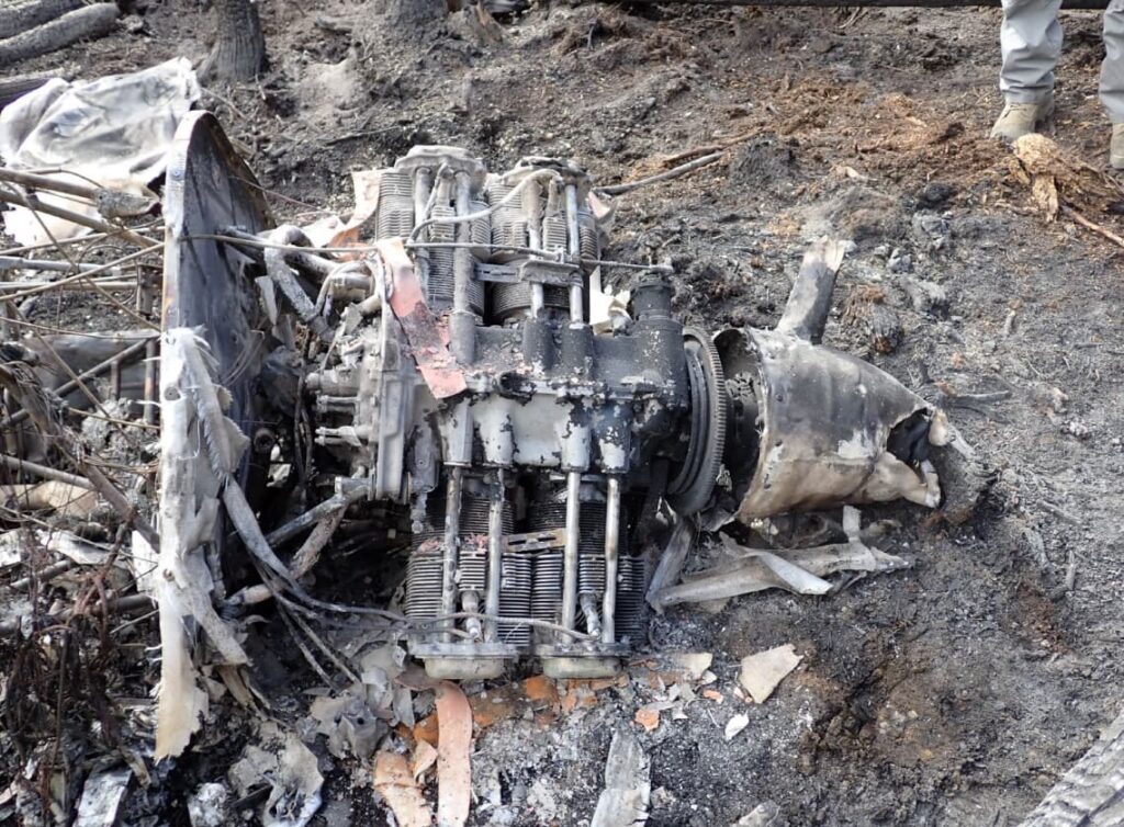 Wreckage of a small aircraft frame in a forest, showing exposed metal tubing with mountainous terrain in the background.
